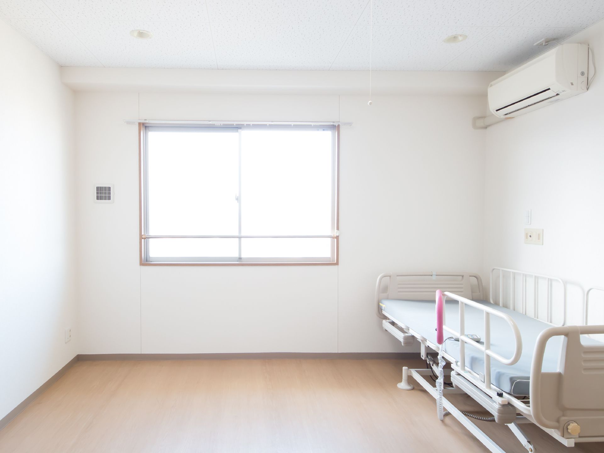クーラーと介護用ベッドを備えた、腰高窓があるフローリング調の個室