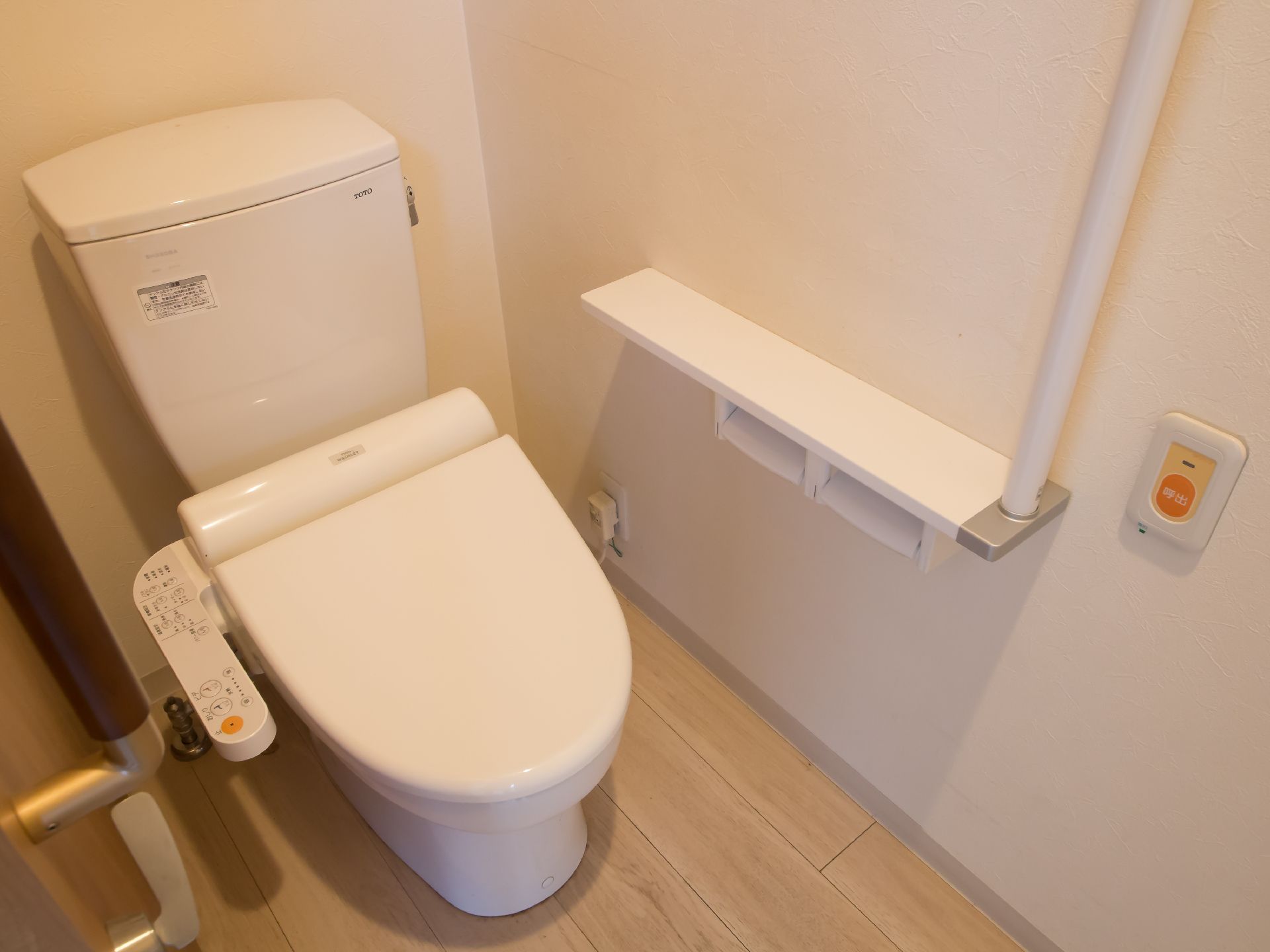 居室のトイレには温水洗浄便座を設置している。便座横に操作パネルがある。壁には手すりと非常ボタンが取りつけられている。