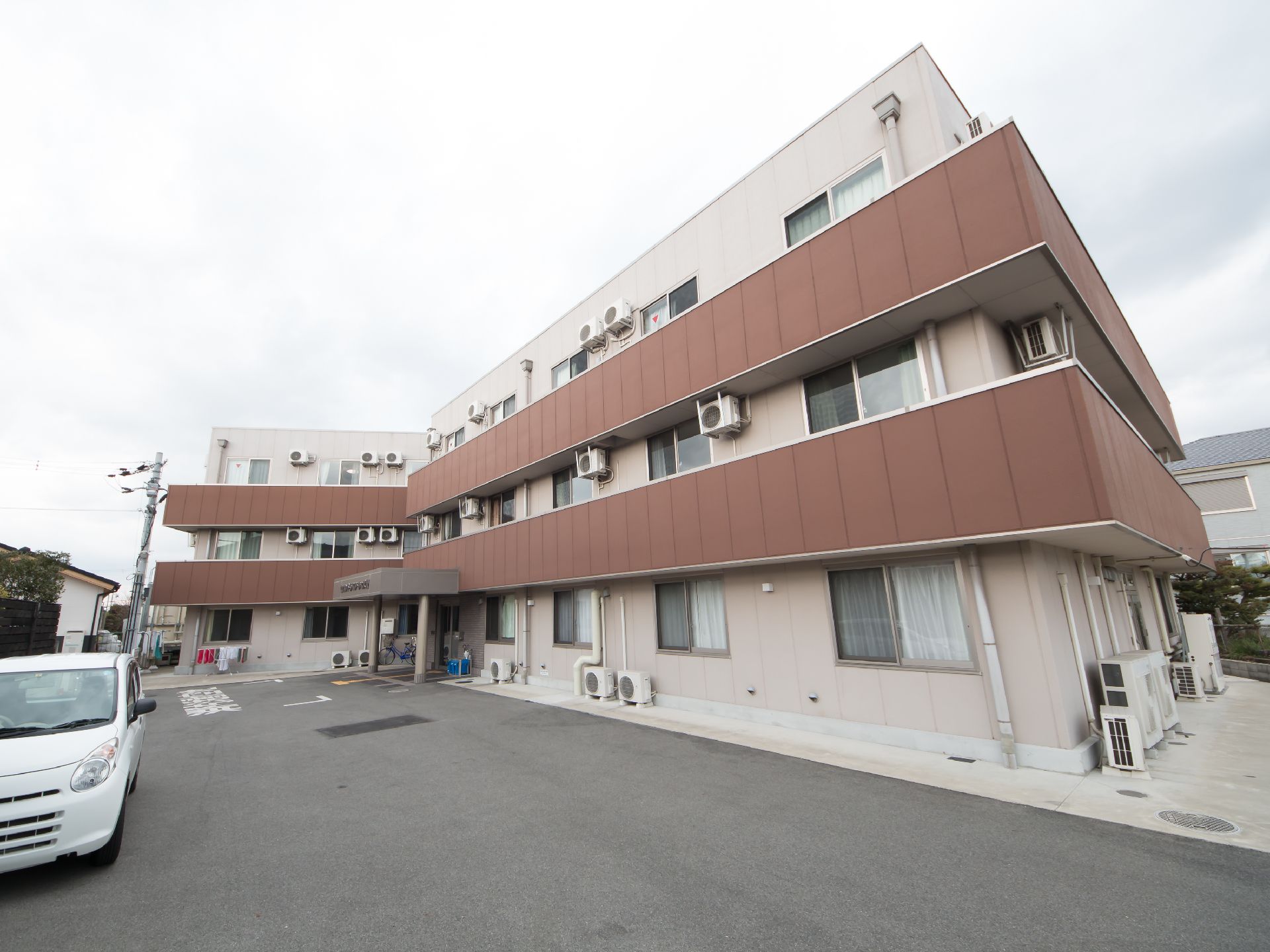白と茶色に塗り分けられた建物で３階建てである。施設の前は駐車スペースになっており、車が１台停められている。