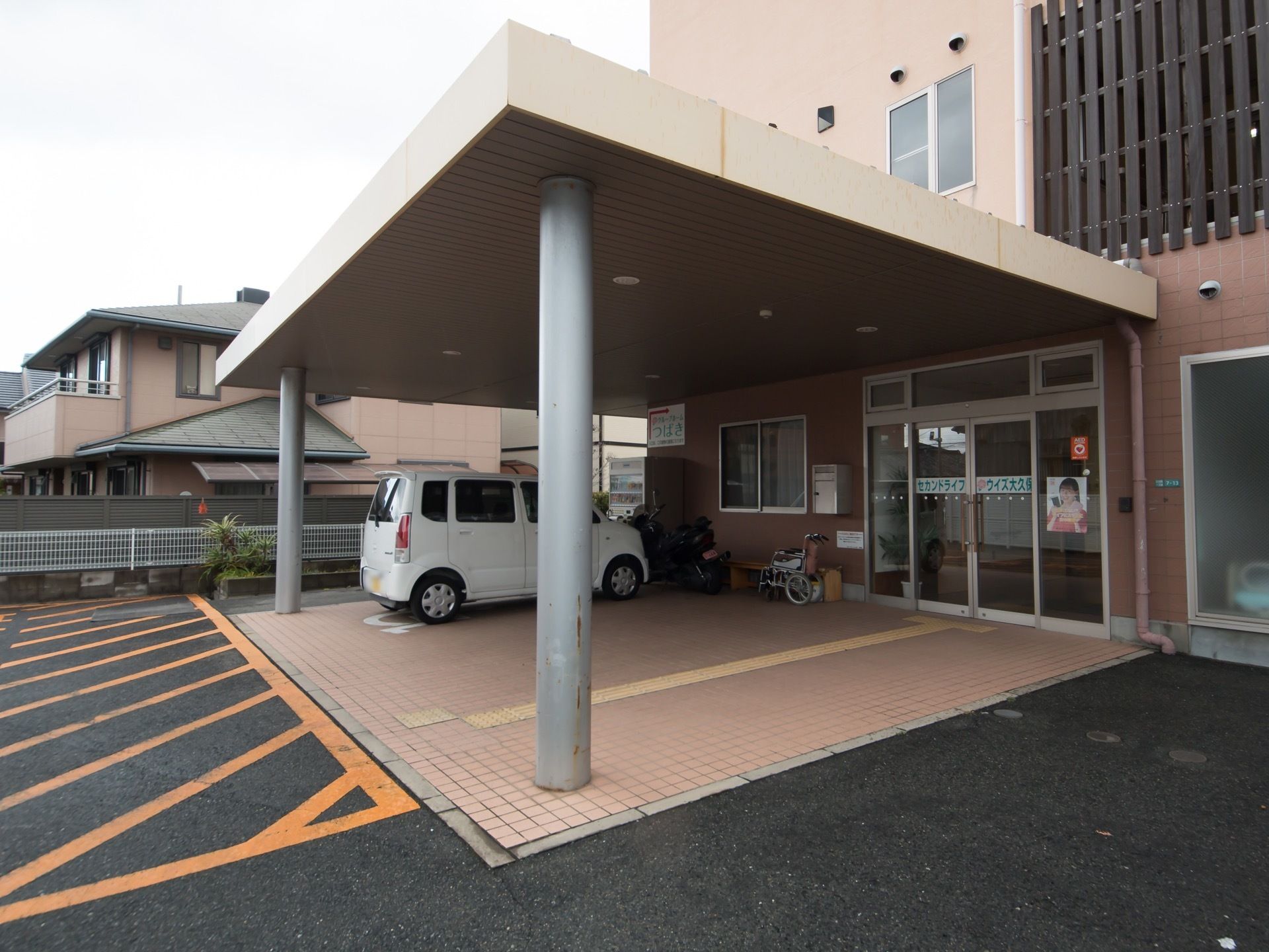 玄関前には大きな屋根が設置され、白い軽自動車が停められている。ガラスの玄関に施設の名称が表示されている。