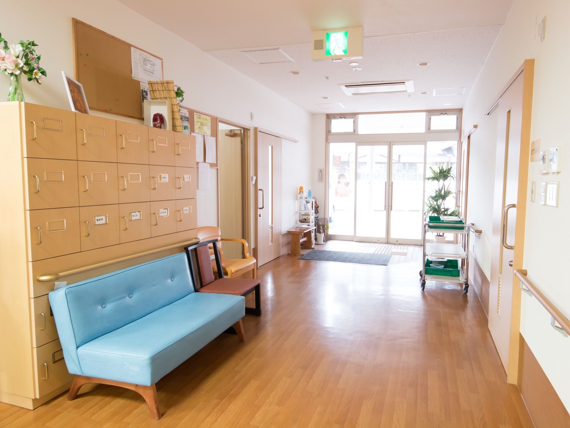 背の高い薄い茶色の靴箱の前に水色のベンチと茶色い１人掛けの椅子が置いてある。玄関前に灰色の玄関マットが敷かれている。