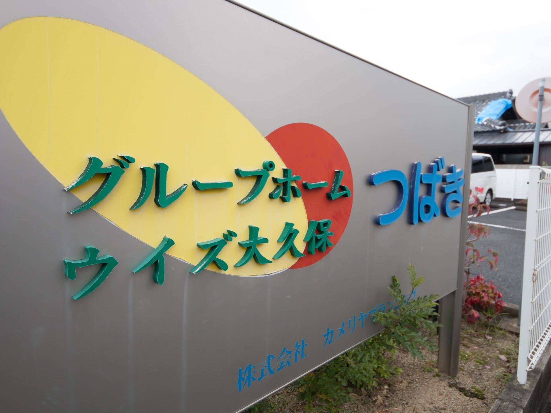 銀色のパネルに緑と水色の文字で施設の名称が表示されている。緑色の文字の下に赤と黄色い楕円が描かれている。