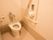 蓋のないタンクレスの白い洋式トイレがある。壁に手すりと洗浄センサーとペーパーホルダーが設置されている。