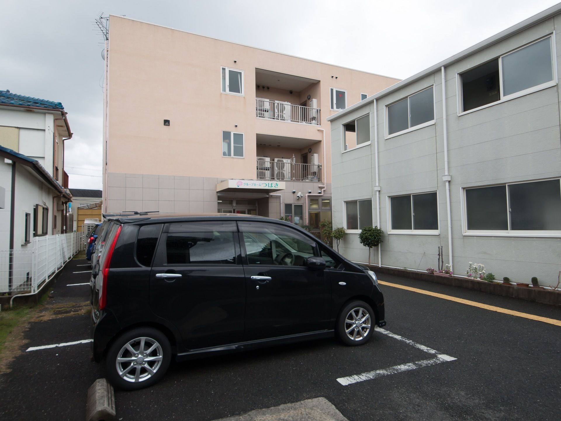 ベージュ系の色を基調とした外壁の３階建ての施設である。玄関前の敷地内に多くの駐車スペースが確保されている。