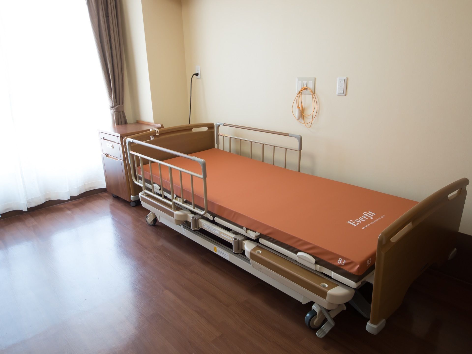 高さのある窓が設置されている居室には、キャスター付き収納棚と介護用ベッドが配置され、壁には緊急呼び出しブザーが取りつけられている。