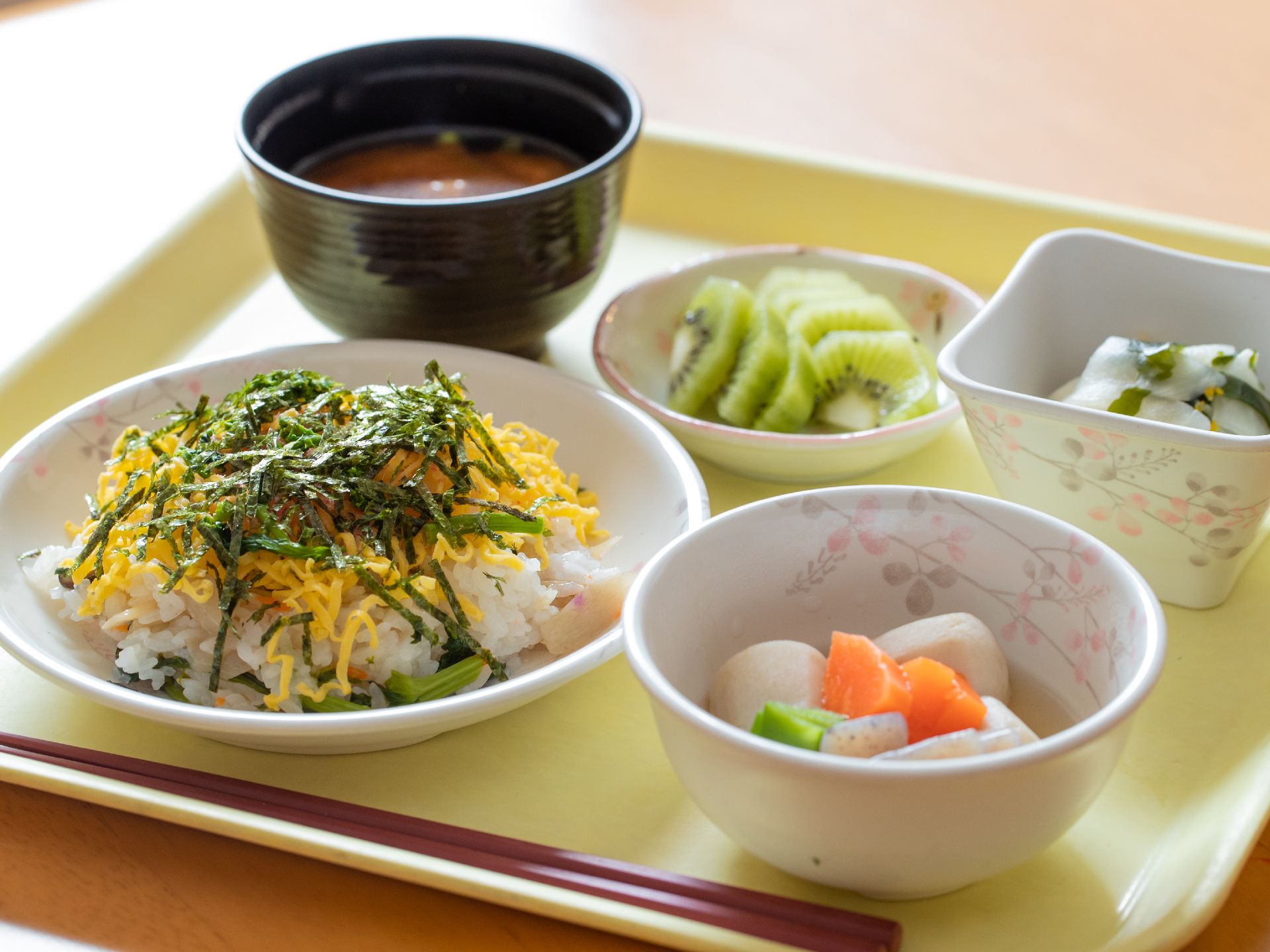 ご飯、味噌汁と里芋の煮物や漬物、果物が用意されている。白米の上には錦糸卵が乗っていて、彩りがきれいである。