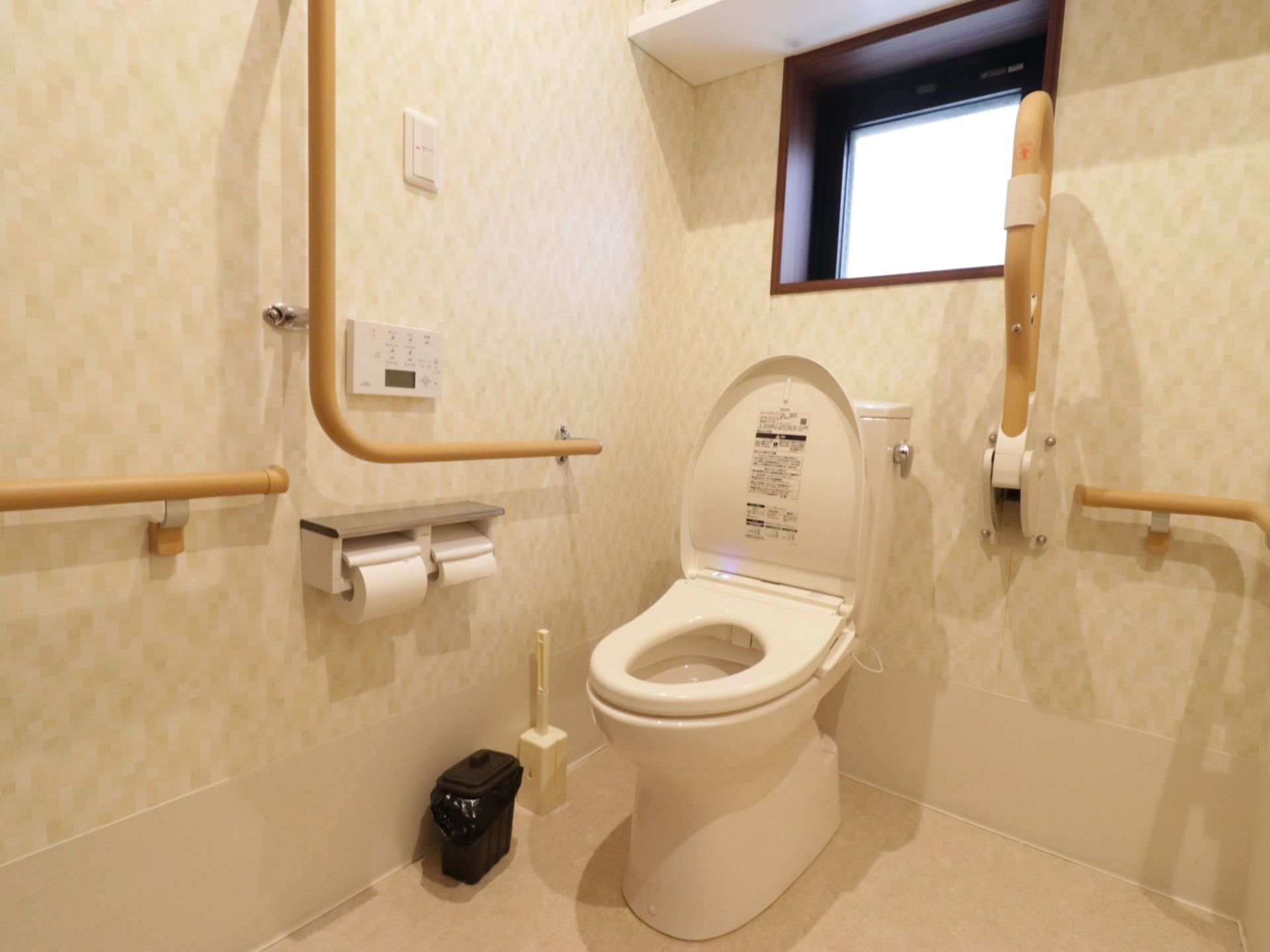 優しい色合いの壁紙が貼られていて、落ち着けるトイレだ。温水洗浄便座が設置されている。壁にはいくつも手すりがついている。