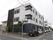 角地に建つ施設は、モノトーンを基調とした洗練されたデザインが特徴の３階建てである。建物とフェンスの間に木が植えられている。