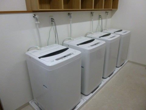 白い床と壁の洗濯室に、防水パンの上に置かれた洗濯機が４つある。洗濯機は縦型で、上には木でできた棚が置いてある。