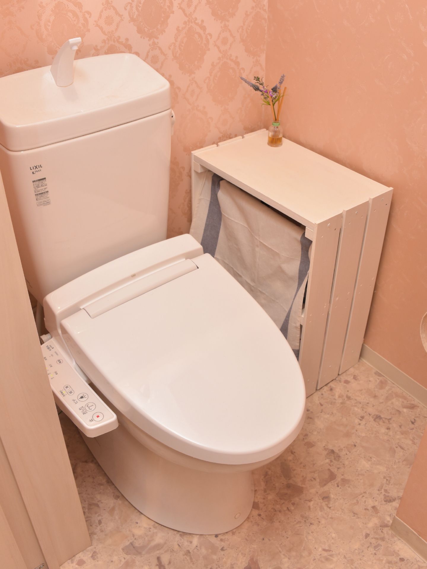 トイレの壁紙はピンク色で温かみのある色合いである。温水洗浄便座なので快適に利用できる。白い棚が置かれており、棚の上にハーブがおいてある。