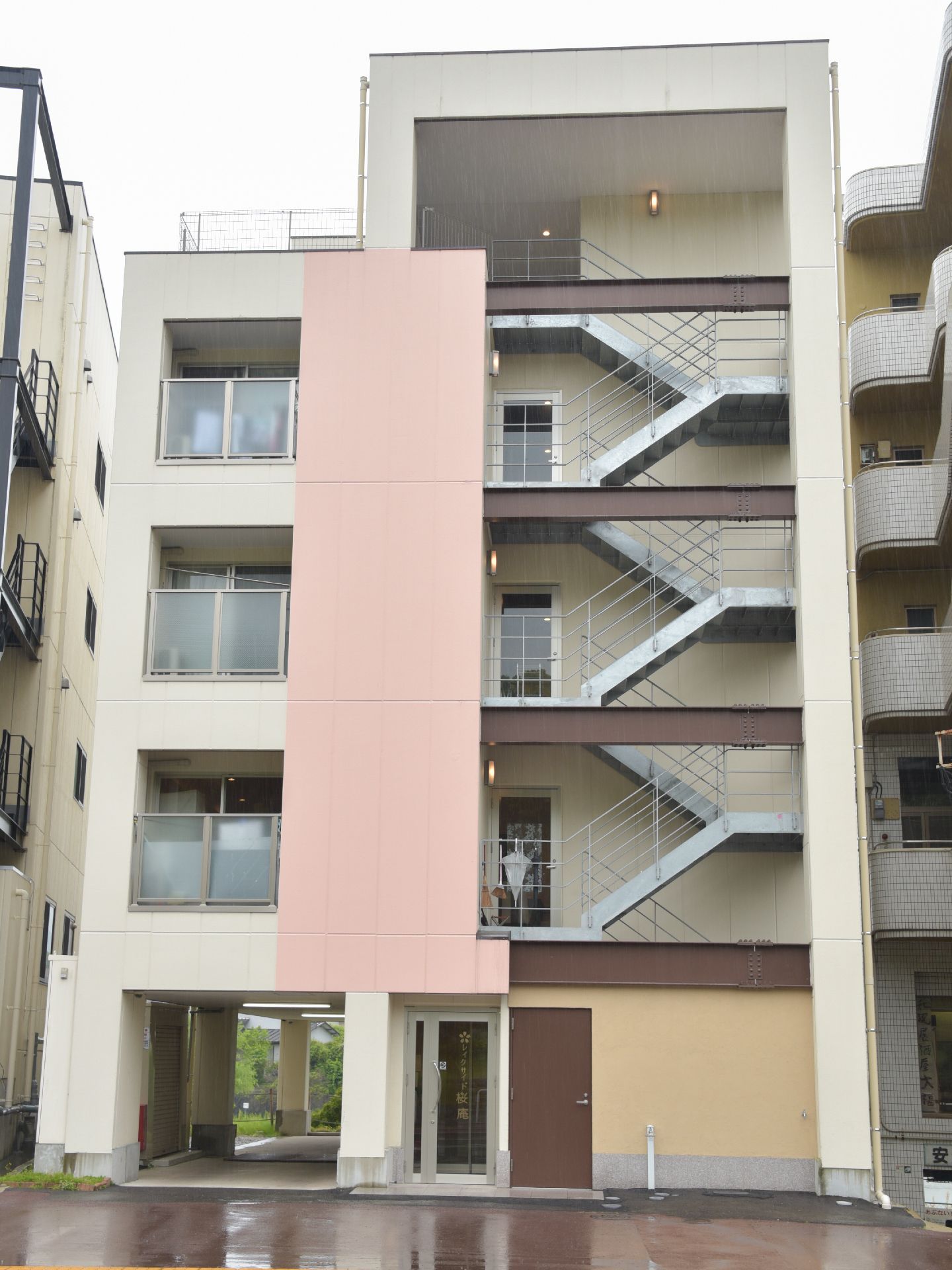 施設は5階建てで、外階段がついている。細長い建物で、白を基調にピンクや茶色などのアクセントがついている。左側に部屋のベランダがある。