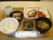木目柄のトレイにのせられた食事の写真である。ご飯、味噌汁と主菜の魚料理のほかに２品が付いた和食メニューである。