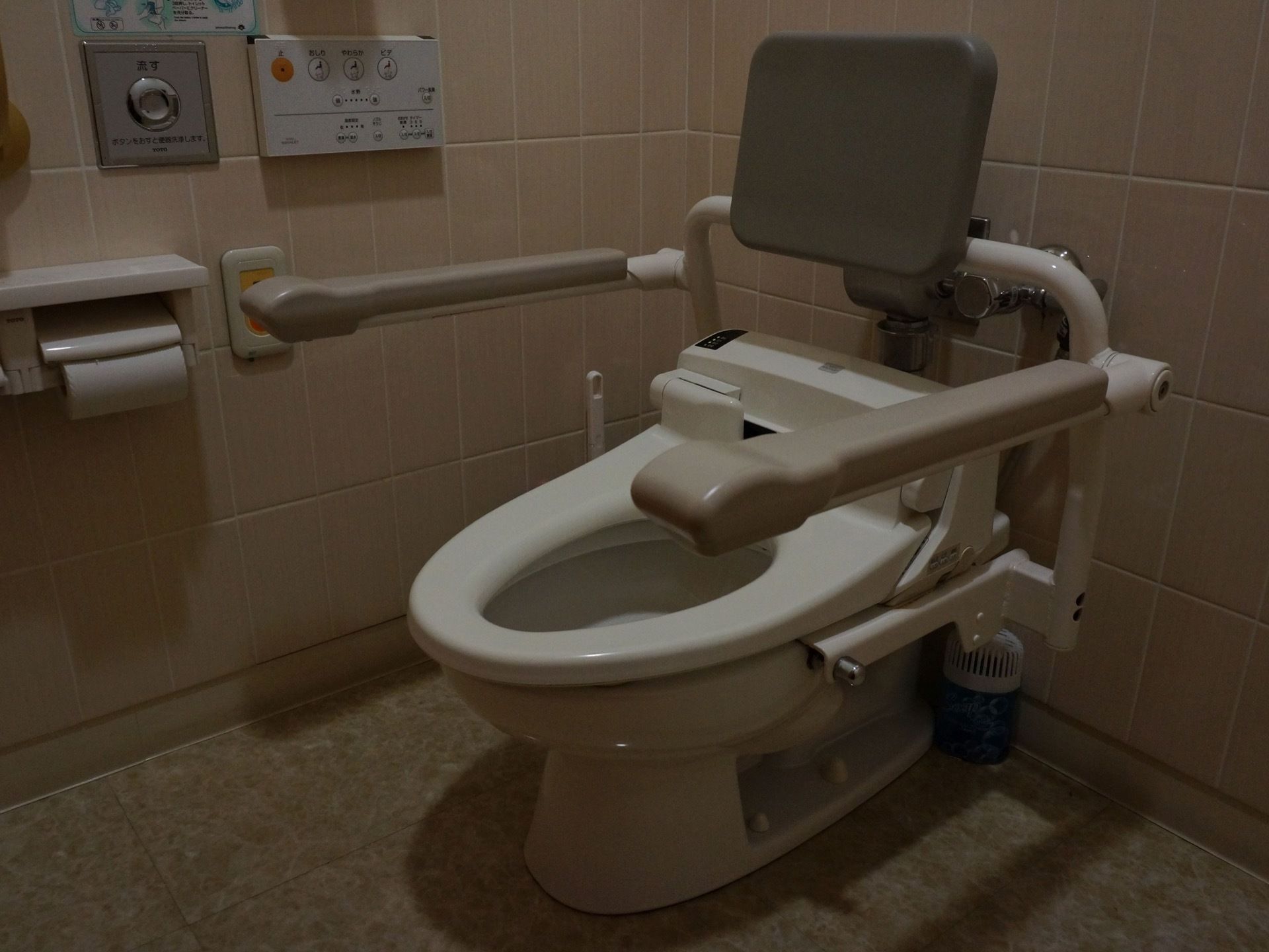 落ち着いた色合いのトイレで、手すりや背もたれが完備されている。壁にリモコン、緊急呼び出しボタンなどがある。