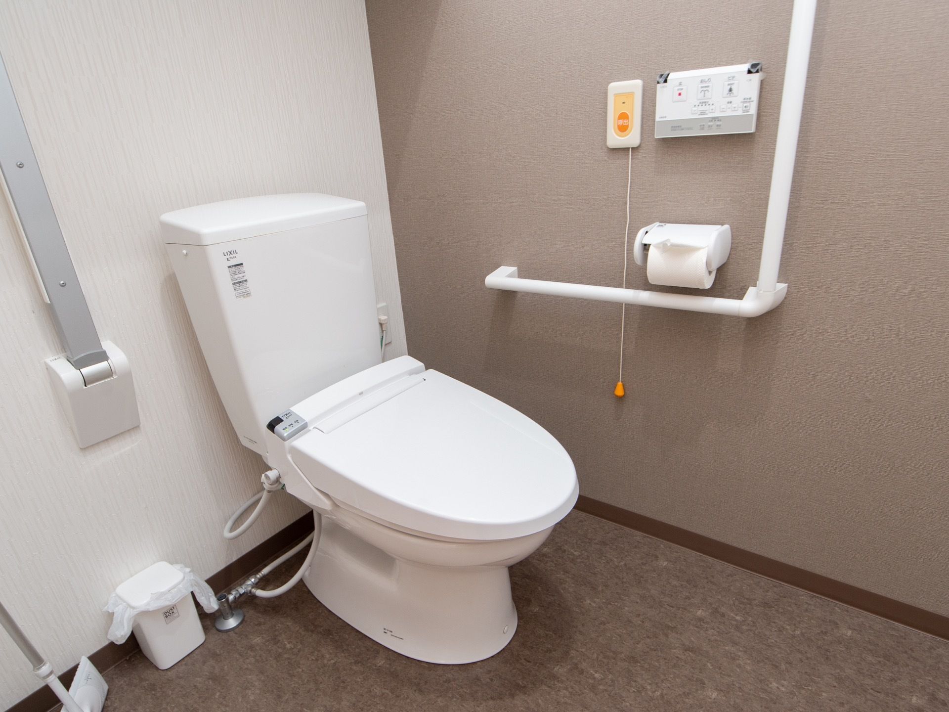 温水洗浄機能付きのトイレが設置されている。グレーの壁にはＬ字型の手すりや操作パネル、ナースコールが取りつけられている。