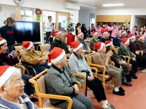 大きな部屋に集まった入居者の皆様が、椅子に座りサンタの帽子をかぶってイベントを楽しむ様子