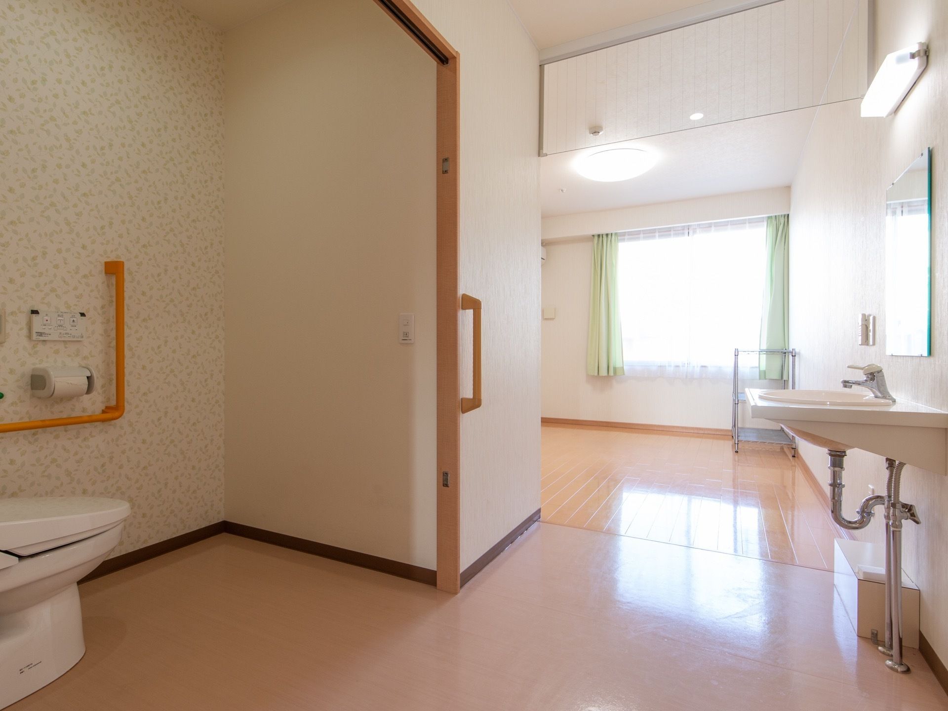 扉から居室内を見た様子である。左側に扉が開かれた状態のトイレが完備され、右側に洗面台、奥には窓が設置されたフリースペースが広がっている。