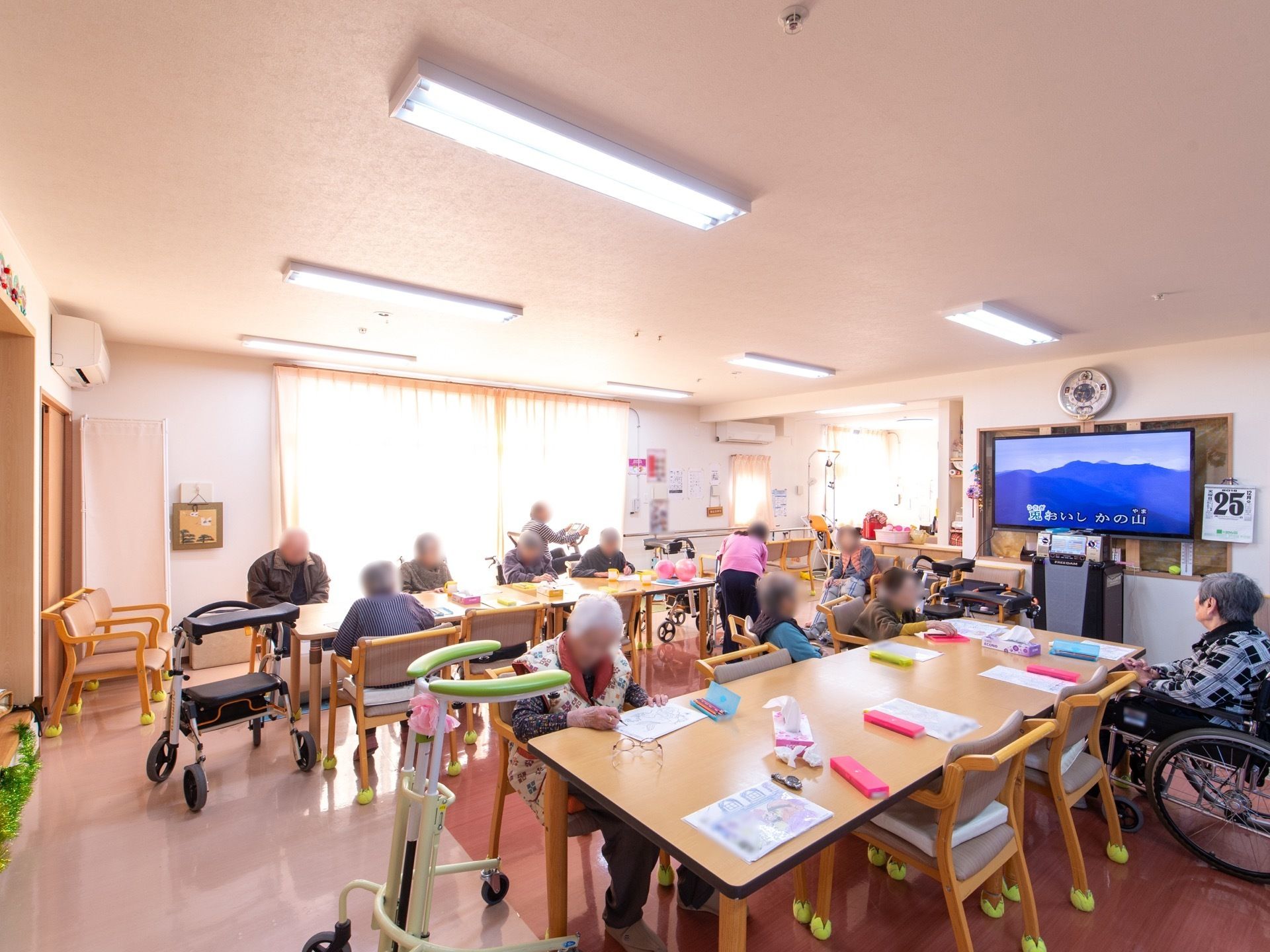 大型テレビが設置された共有スペースである。入居者様皆様が集いレクリエーションを行っている。車椅子や歩行器をご利用の方の姿が見える。