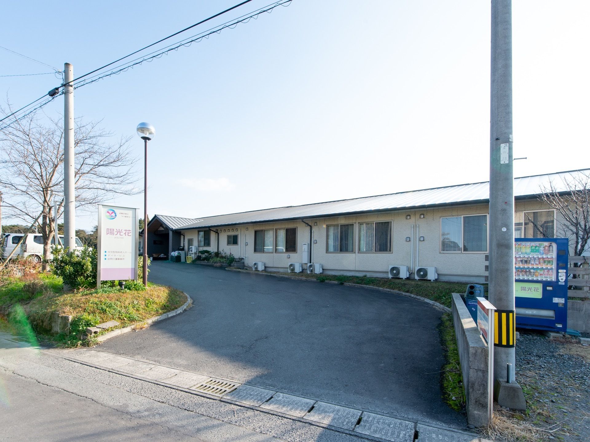 道路から平屋建ての施設入口への道なりである。左側には施設看板が、右側には自動販売機が設置されている。
