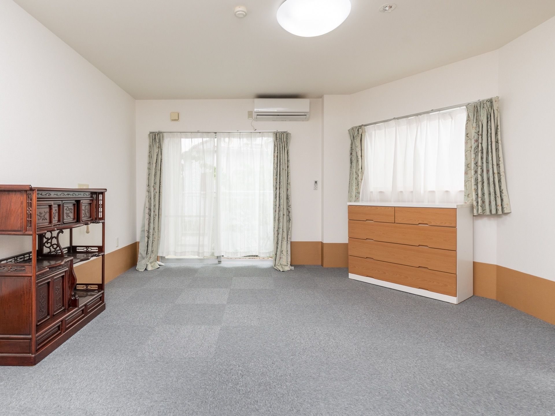 居室は広々としており、2ヶ所の窓にはグレーのカーテンが掛けられている。室内には、収納棚が2つ置かれている。