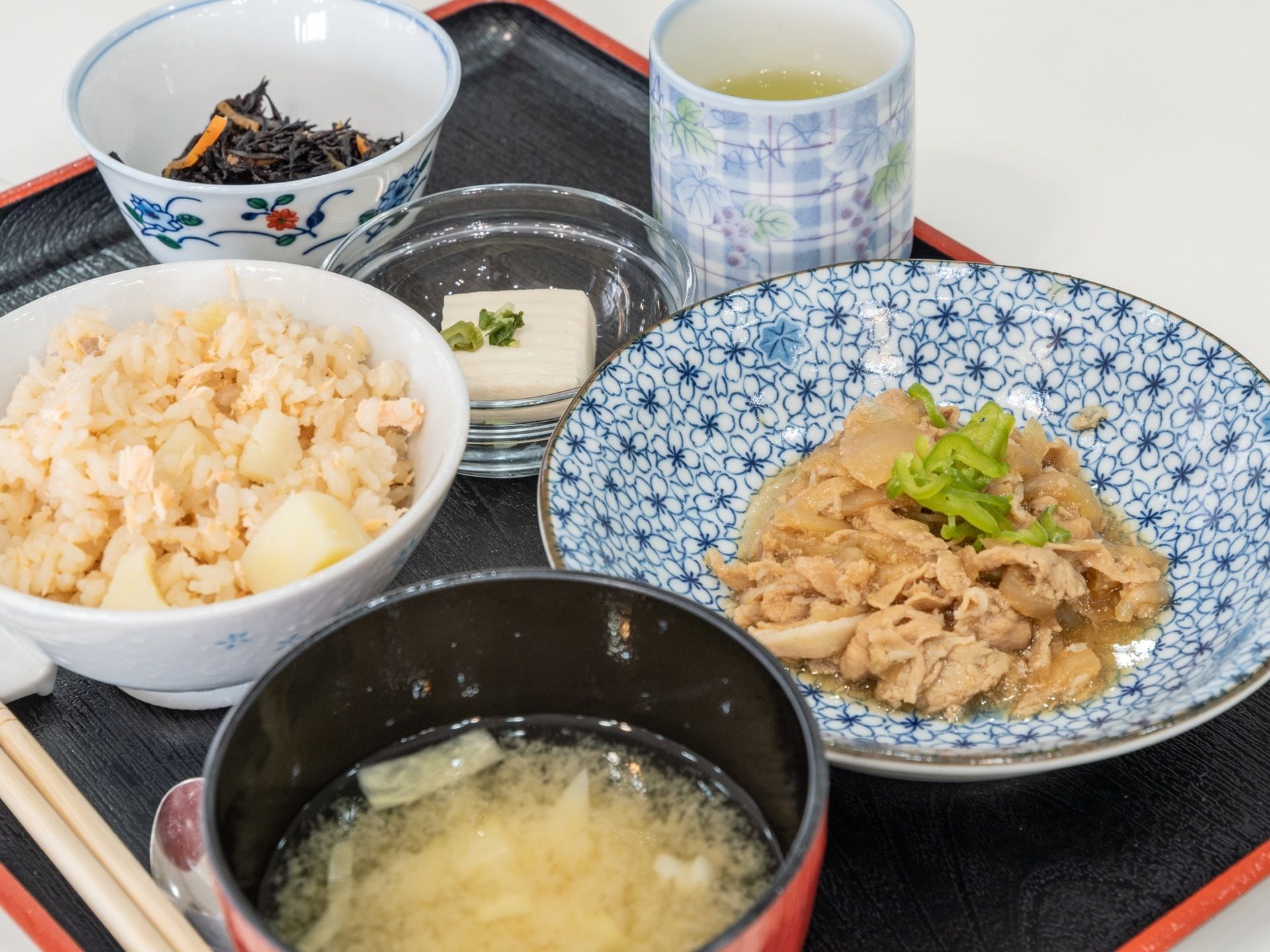 黒いトレーの上に、5つの器に食事が盛りつけられている。一緒に、青色の湯のみに入ったお茶が添えられている。