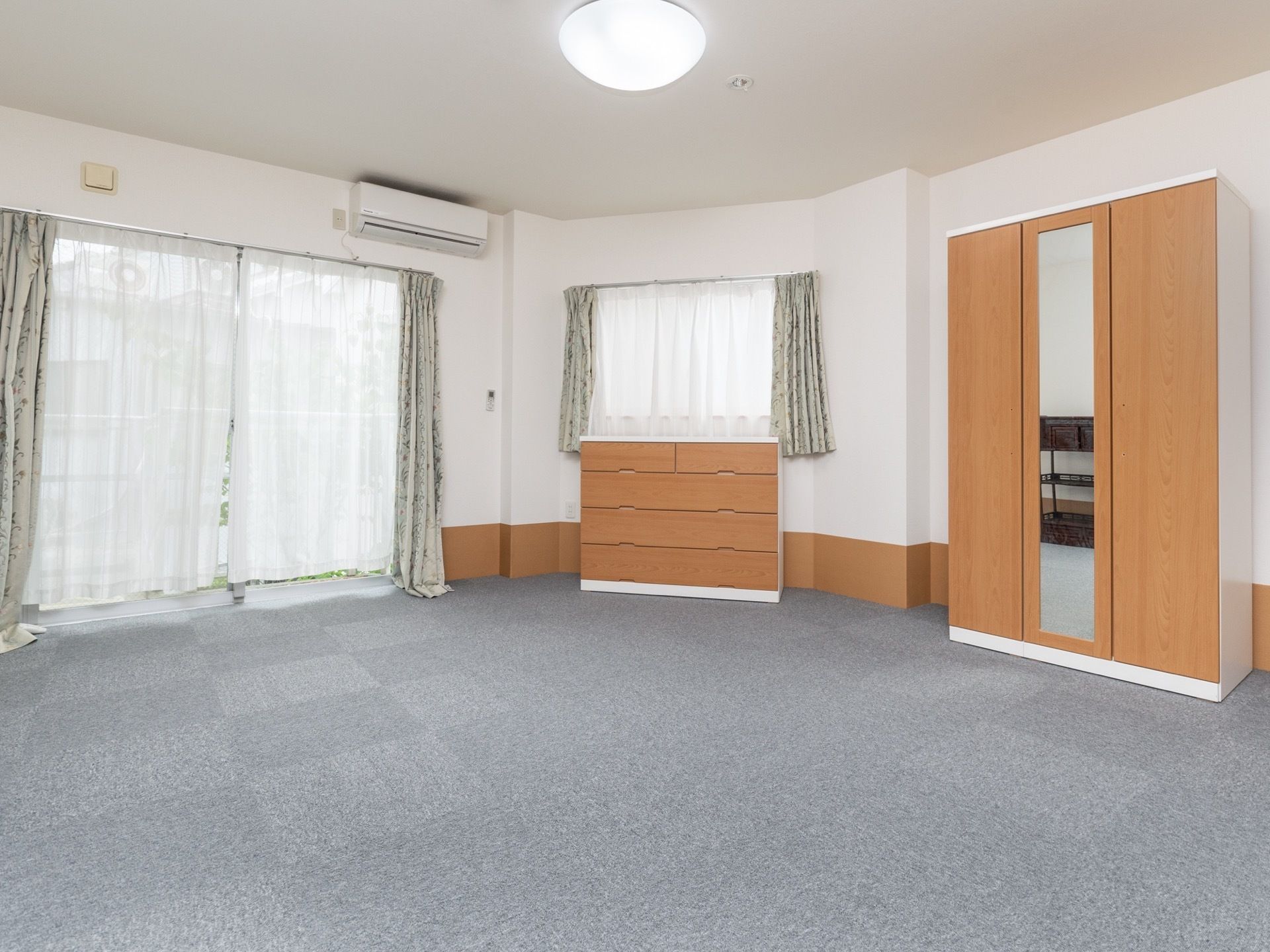 グレーの絨毯が敷かれた広い居室である。窓が二カ所にあり、整理ダンスや洋服ダンスが置かれている。エアコンが設置されている。