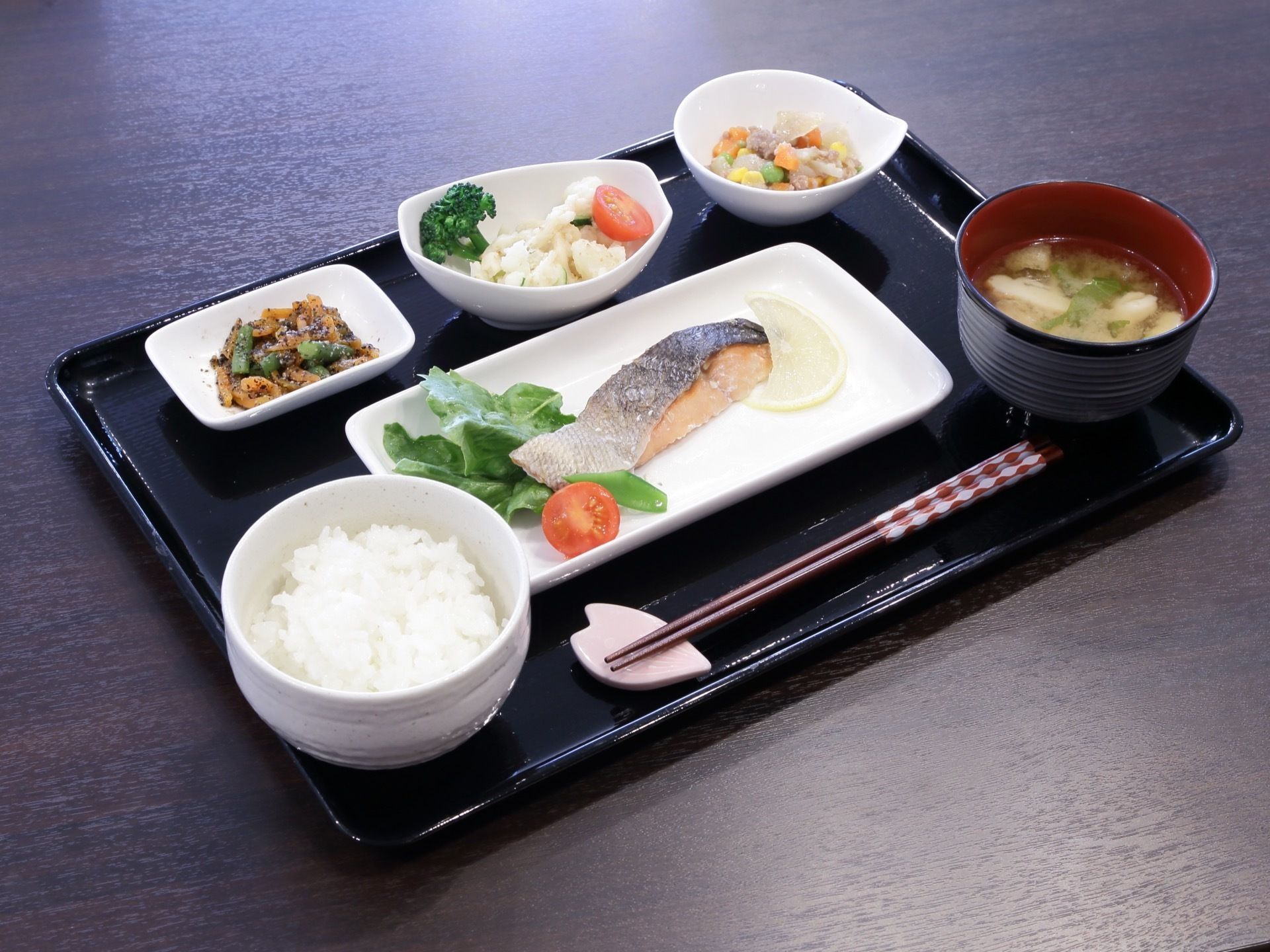 施設にて提供される食事。白米を主食に据えて味噌汁付きの和食メニューで、主菜となる焼き魚が中心に置かれている。