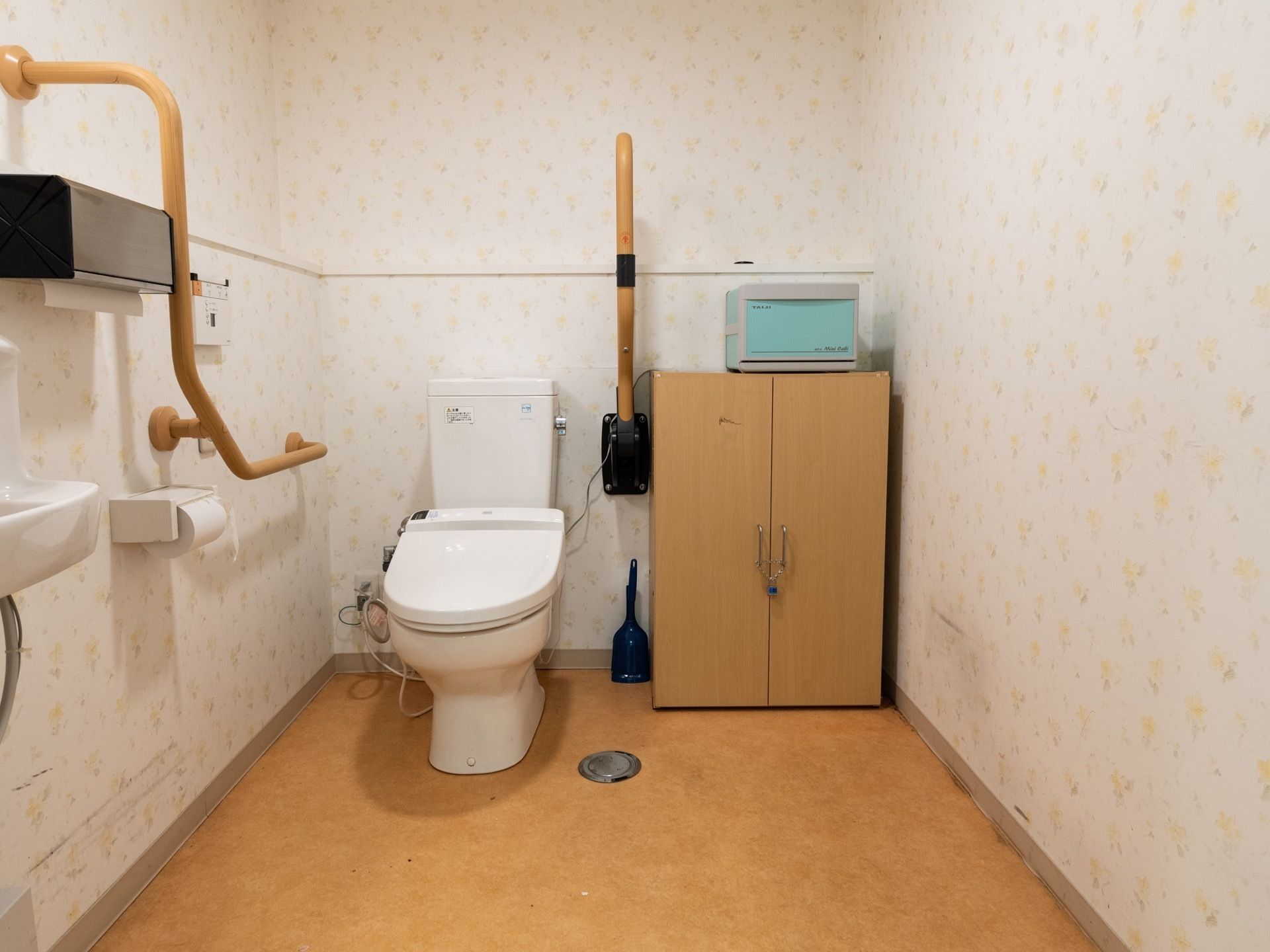 模様入りのクロスを取り入れたトイレスペースである。便器の両側に手すりを設置している。隣に両開きの扉の収納庫を備えている。