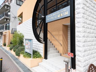 階段のあるエントランスである。出入口上部に横書きの看板があり、建物傍に緑豊かな花壇を設けている。階段の横側に手すりを設置している。