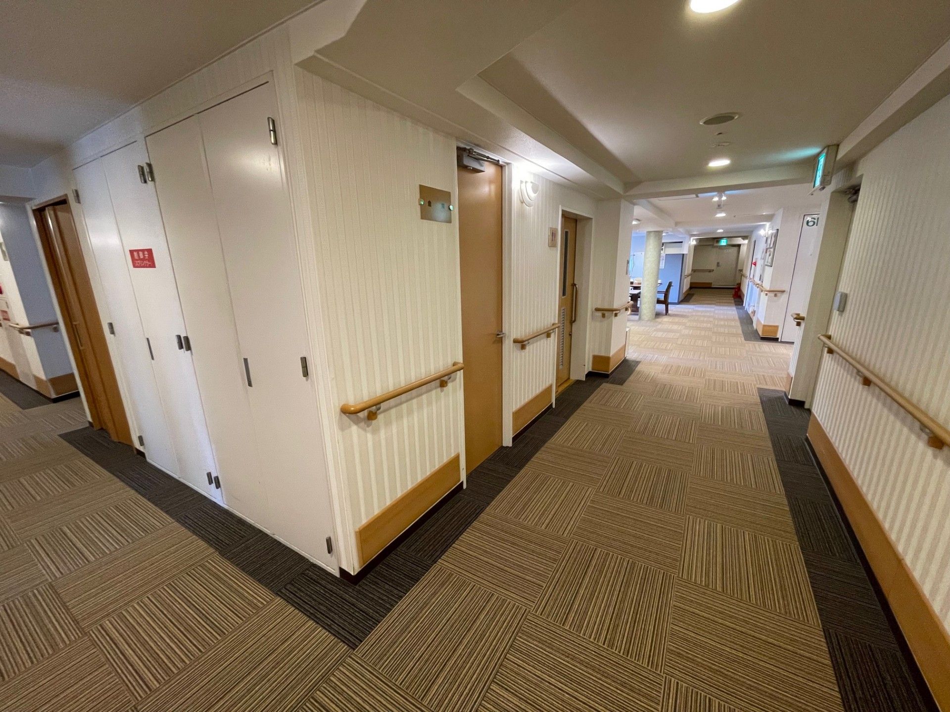 ストライプの壁、模様入りのフローリングの通路は、お洒落な雰囲気である。壁に手すりを完備し、壁際に掃除道具のワゴンがある。