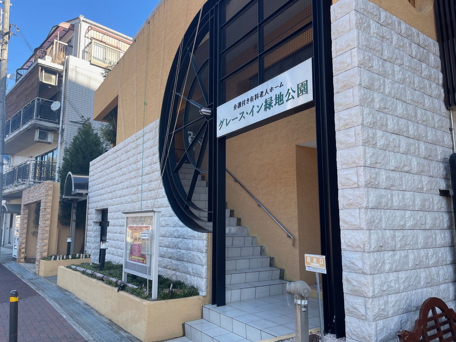階段のあるエントランスである。出入口上部に横書きの看板があり、建物傍に緑豊かな花壇を設けている。階段の横側に手すりを設置している。