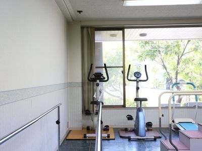 大きな窓がある明るい空間のトレーニングルームには、さまざまな機能訓練機器を備えている。ガラス戸の向こうには、緑豊かな風景が広がっている。