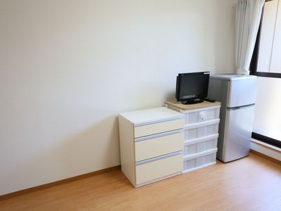 お部屋の隅に小さめの冷蔵庫と収納ボックスを並べて配置し、上にテレビを置いている。横側にガラス戸があり、カーテンを掛けている。
