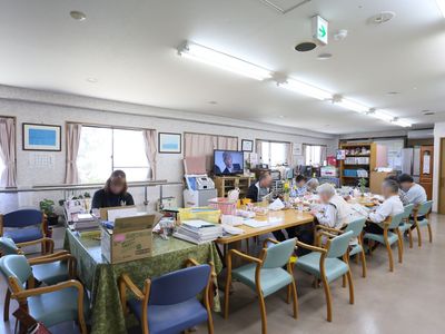 縦に長い共有スペースにテーブルを向かい合わせに並べている。横側に窓が並びテレビを置いている。天井に細長い２列の照明を５ヶ所設置している。