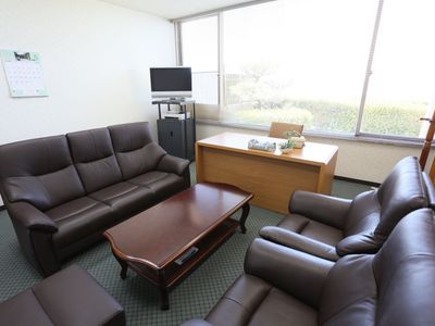 大きな窓際にデスクを置き、中央にテーブルとそれを囲むようにソファを配置している。角にテレビを備え、壁にカレンダーを掛けている。