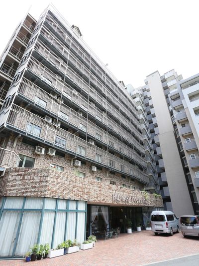 高層の建物で、玄関前は広々としていて屋根が取りつけられている。カフェ風のテーブルと椅子が置かれている。