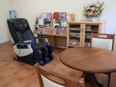 丸テーブルと椅子のセット、マッサージチェア、本棚、マガジンラックが置かれている。奥にはウォーターサーバーがある。