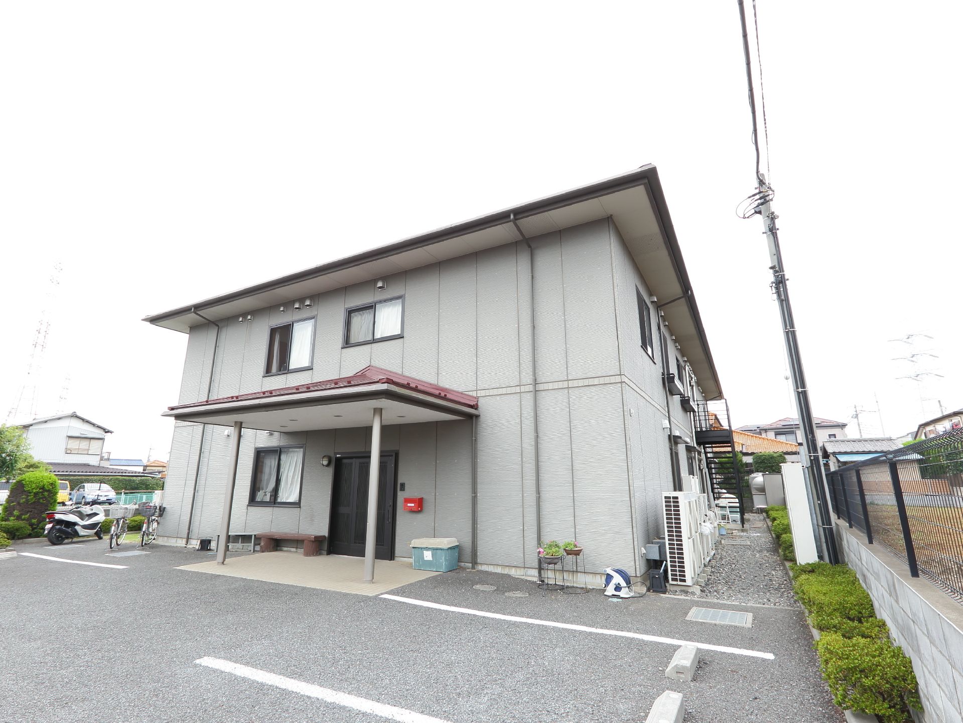 建物の正面はアスファルトの駐車場になっていて、向かって右側は砂を敷いている。エアコンの室外機が複数台ある。隣とは柵で区切っている。