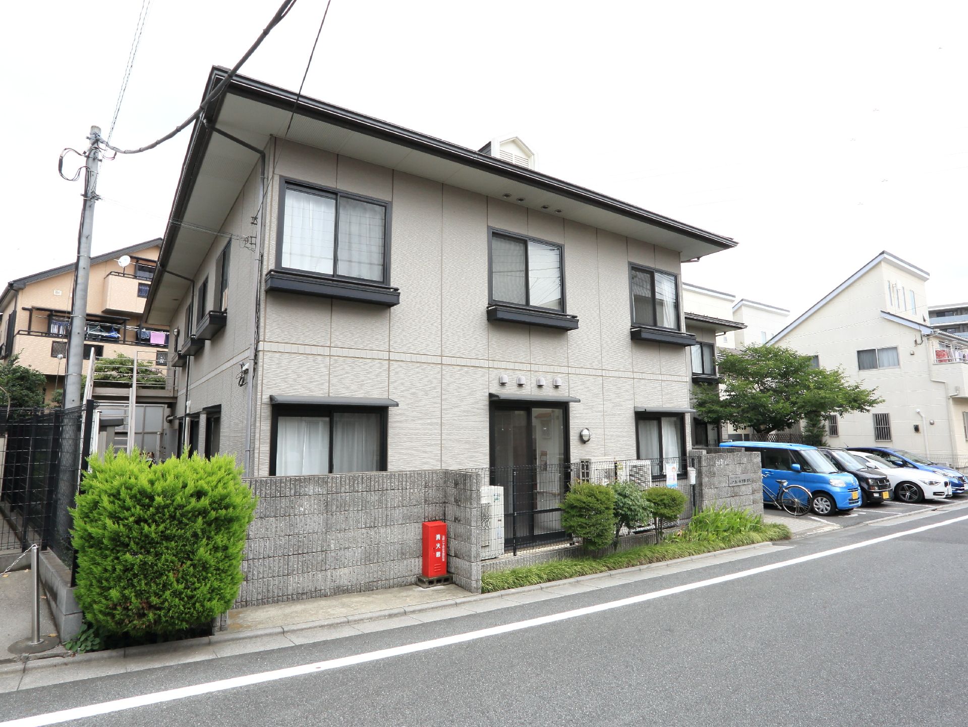 施設前の公道と建物の間にはコンクリートフェンスがあり、道路に面した外側には小さな植え込みと、消火器の入った赤いボックスがある。