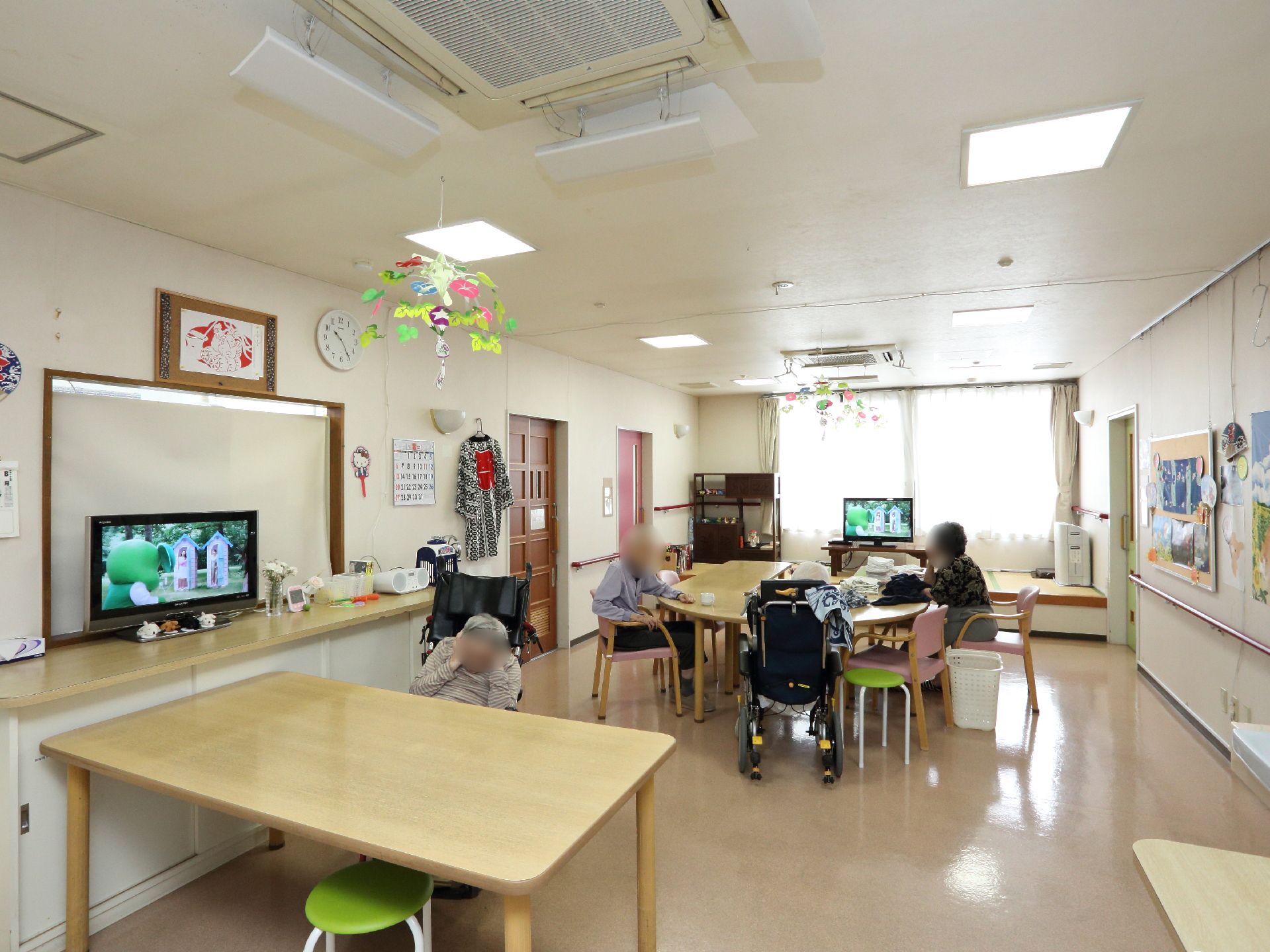 多くの入居者が思い思いの時間や共同作業などに利用することが出来る広い共有空間。壁には掲示物、棚にはテレビなどにぎやかな室内。