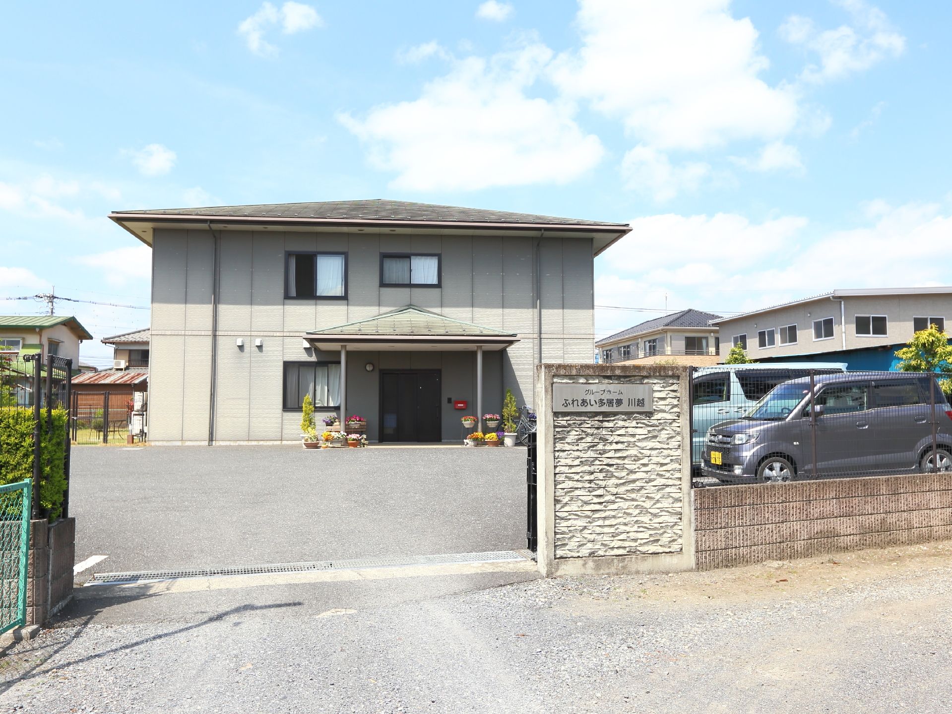 敷地はフェンスで区切られ、門には横書きで施設名が掲示されている。門を入ると駐車場の先に出入り口があり、庇の柱の周りに植木鉢が置いてある。