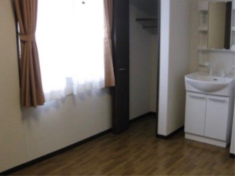 居室には白い化粧洗面台が設置されていて、その隣にクローゼットがある。床はフローリングで、窓にはレースとベージュのカーテンが掛かっている。