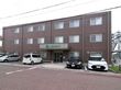 こげ茶色の外壁には、オレンジ色の細いラインが2本入っている。建物の手前は駐車場で、白や黒の数台の車が置かれている。