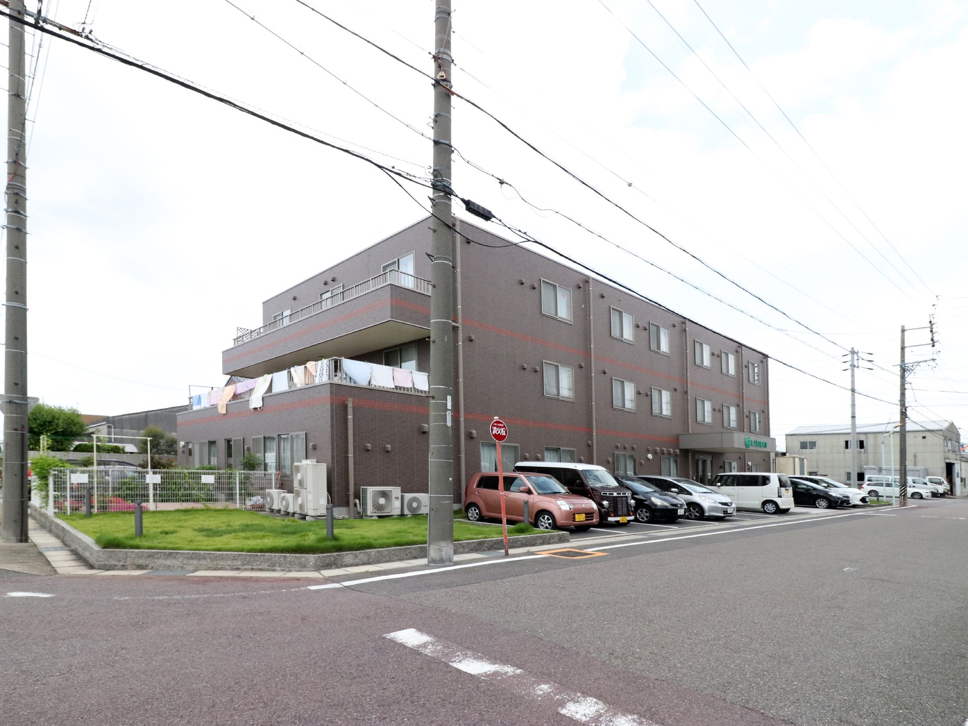 大きな道路の角地に建っている施設は3階建てである。前方の駐車スペースには、オレンジ色の軽自動車など7台の車がある。