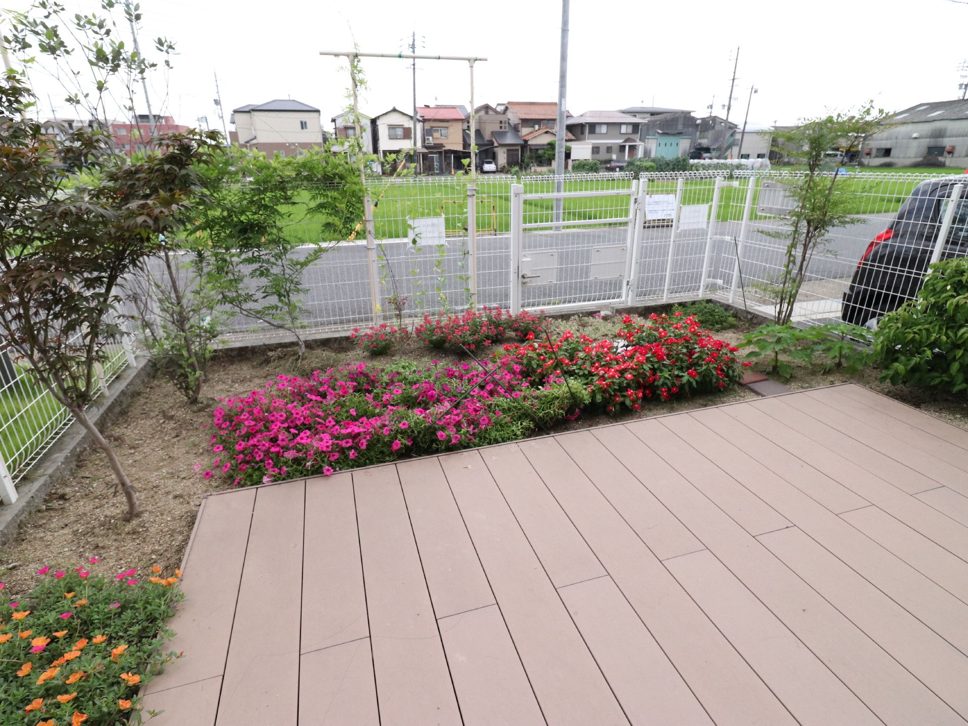 ピンク色のペチュニアの花などが、数多く咲いている。その横には低木もある。バルコニーの周囲には、白い柵が設置されている。
