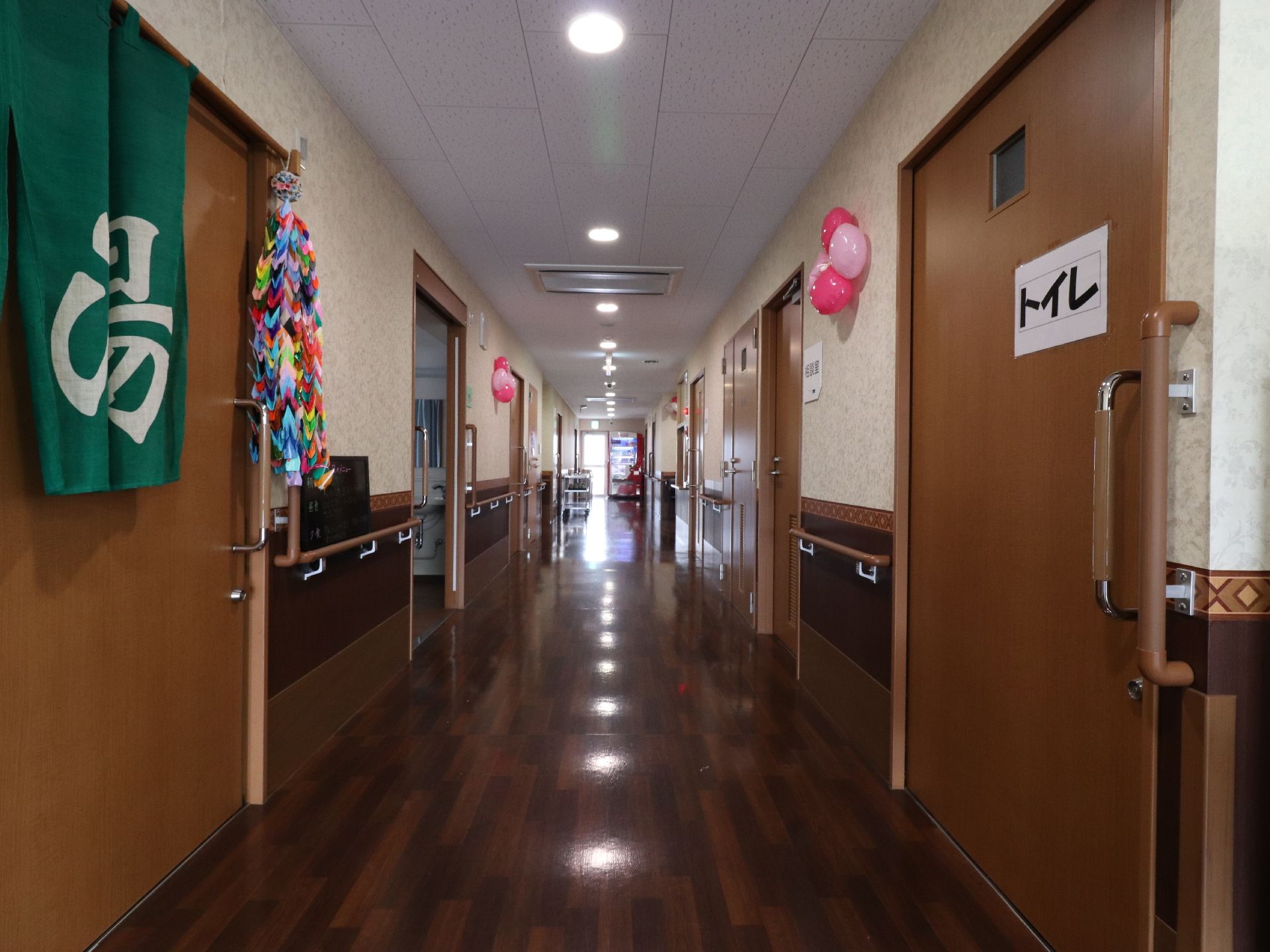 茶色の引き戸が、廊下の両側に並んでいる。左側にはグリーンの暖簾があり、大きな文字で湯と書いてある。千羽鶴も飾ってある。