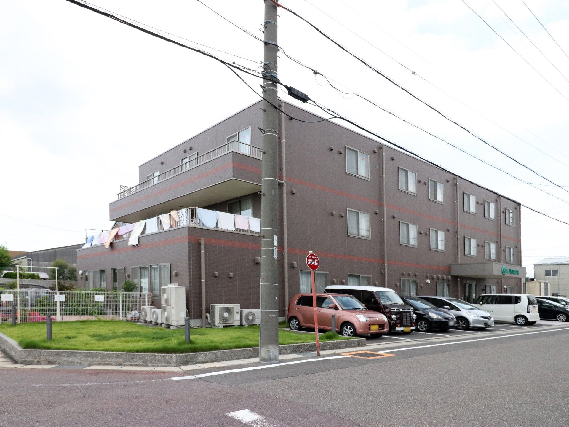 大きな道路の角地に建っている施設は3階建てである。前方の駐車スペースには、オレンジ色の軽自動車など7台の車がある。