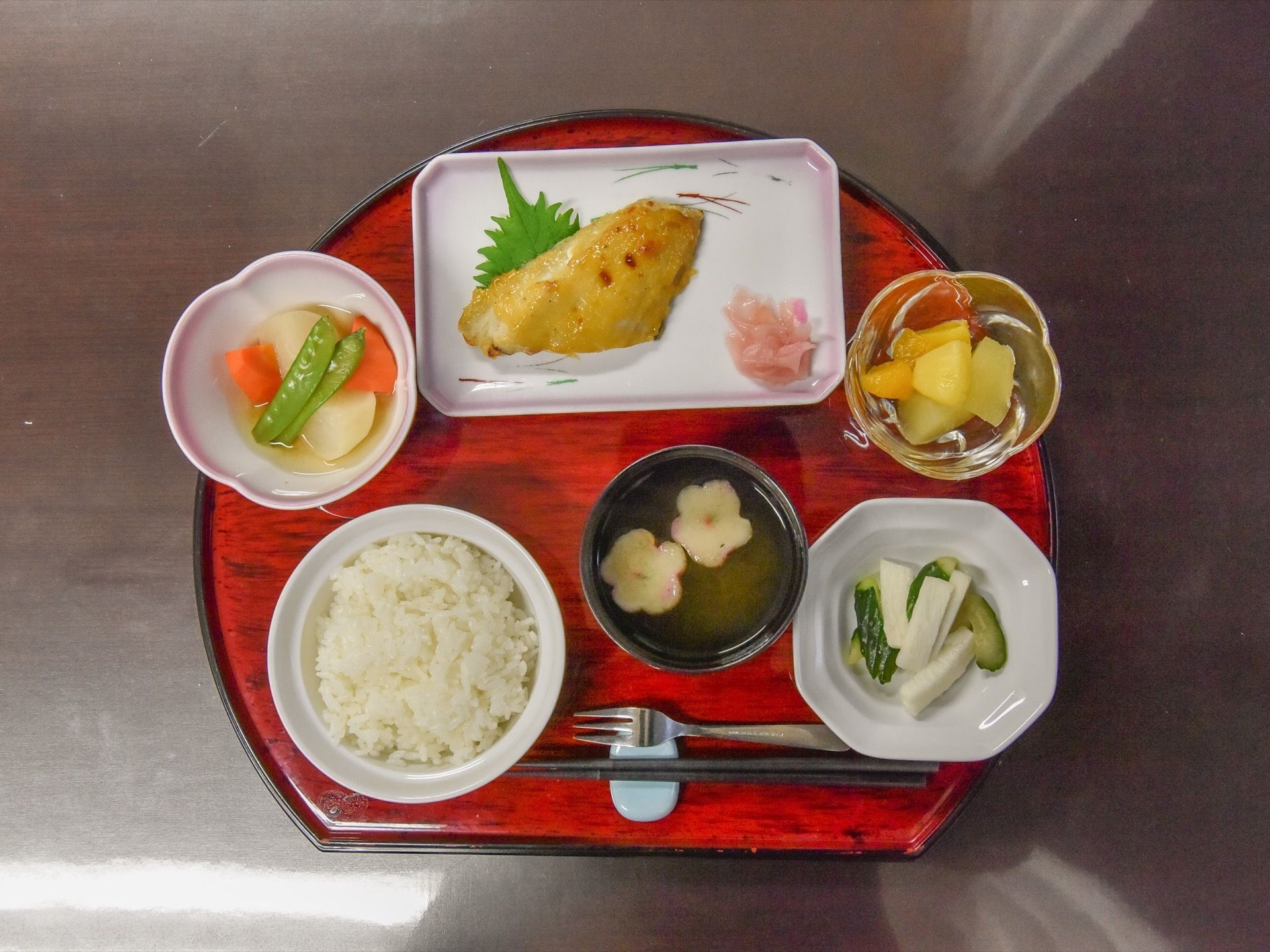 扇形の赤いお盆の上にさまざまな料理がのっている。全部で７種類あり、手前には箸とフォークが添えられている。