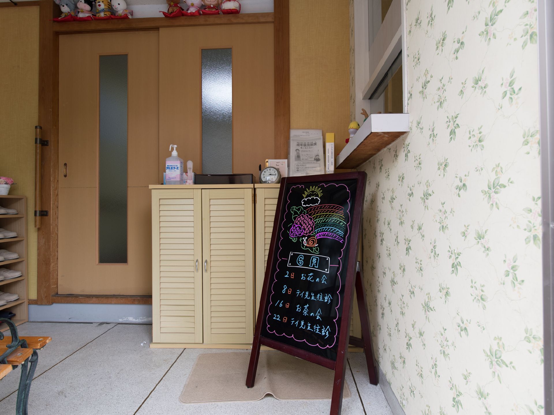 施設のイベントを知らせるサインボードがたっており後ろに戸棚とスライド式のドアの入り口がみえる。右側は受付になっている。