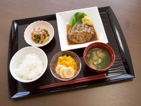 黒いトレーの上に並べられた１食分の食事。ご飯と汁物、メインのハンバーグの他に、副菜の小鉢とデザートが置かれている。