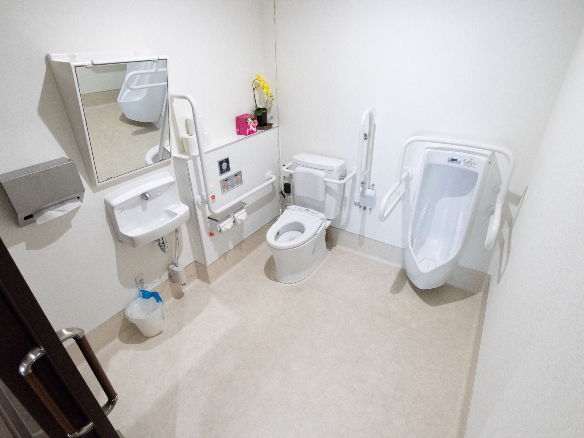 多目的トイレは白で統一された広々とした空間で、手すりが各所に取りつけられている。手前に手洗い器と鏡が設置されている。
